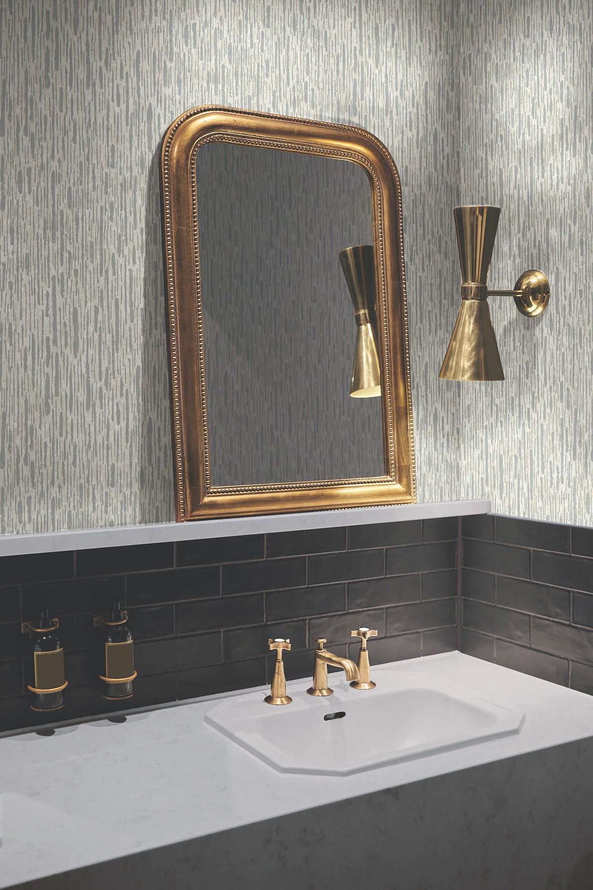 A bathroom featuring a gold-framed mirror, gold faucet fixtures, a gold wall sconce, black tile backsplash, and York Wallcoverings’ Metallic Cascade Wallpaper above a white countertop sink.