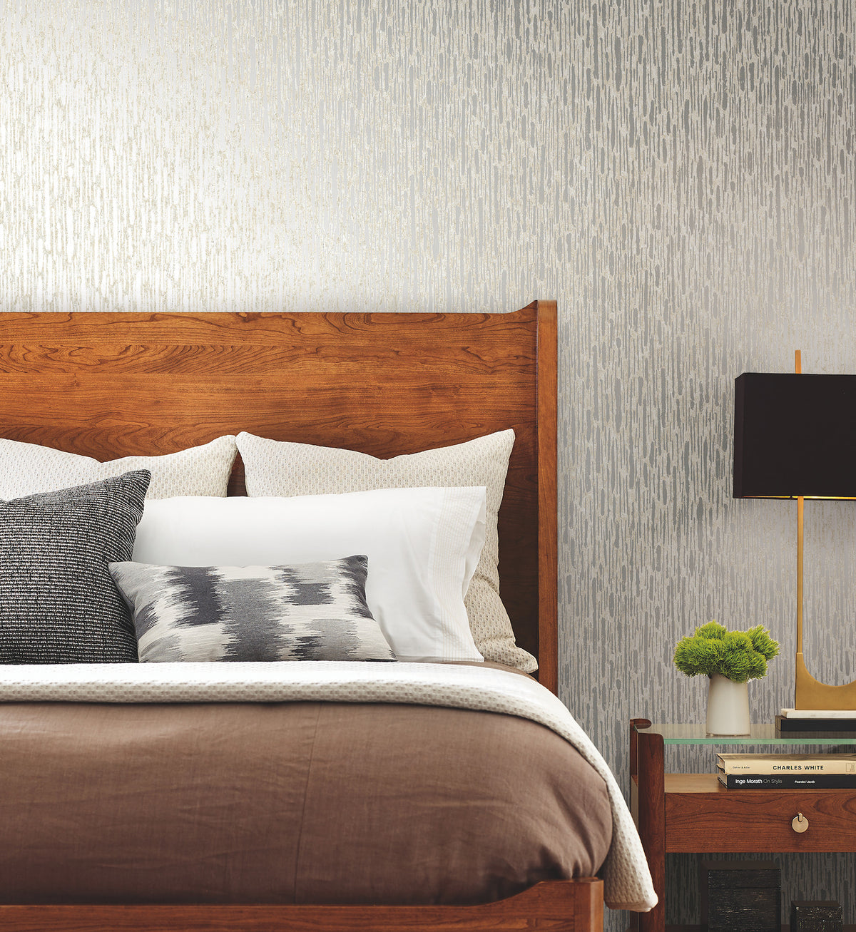 A modern bedroom featuring a wooden bed with neatly arranged pillows and blankets, a nightstand with a lamp, a small plant, and books, set against a gray Metallic Cascade Wallpaper by York Wallcoverings.