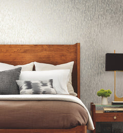 A modern bedroom featuring a wooden bed with neatly arranged pillows and blankets, a nightstand with a lamp, a small plant, and books, set against a gray Metallic Cascade Wallpaper by York Wallcoverings.