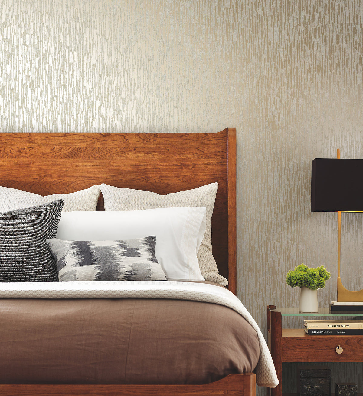 A neatly made bed with a wooden headboard, layered pillows, brown bedding, and a nightstand holding a black lamp, a small plant, and stacked books sits against a wall adorned with Metallic Cascade Wallpaper by York Wallcoverings.
