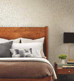 A neatly made bed with a wooden headboard, layered pillows, brown bedding, and a nightstand holding a black lamp, a small plant, and stacked books sits against a wall adorned with Metallic Cascade Wallpaper by York Wallcoverings.
