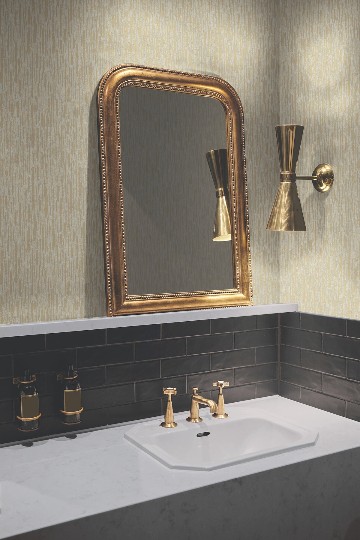 A bathroom sink with gold faucets, a gold-framed mirror, and a gold wall sconce sits against York Wallcoverings’ Metallic Cascade Wallpaper above dark subway tiles and a white countertop.