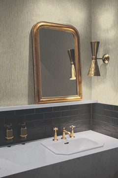 A bathroom sink with gold faucets, a gold-framed mirror, and a gold wall sconce complements a white countertop and dark tile backsplash, while York Wallcoverings’ Metallic Cascade Wallpaper in a light, textured pattern adds an elegant touch.
