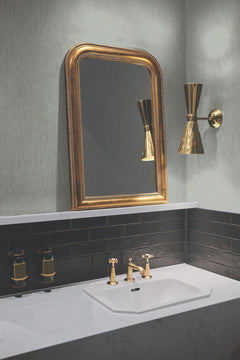 A bathroom sink with a gold faucet sits beneath a gold-framed mirror, with a gold wall light to the right and black tiles below. The gray textured wall above the tiles features Metallic Cascade Wallpaper by York Wallcoverings, and soap dispensers are mounted on the left.