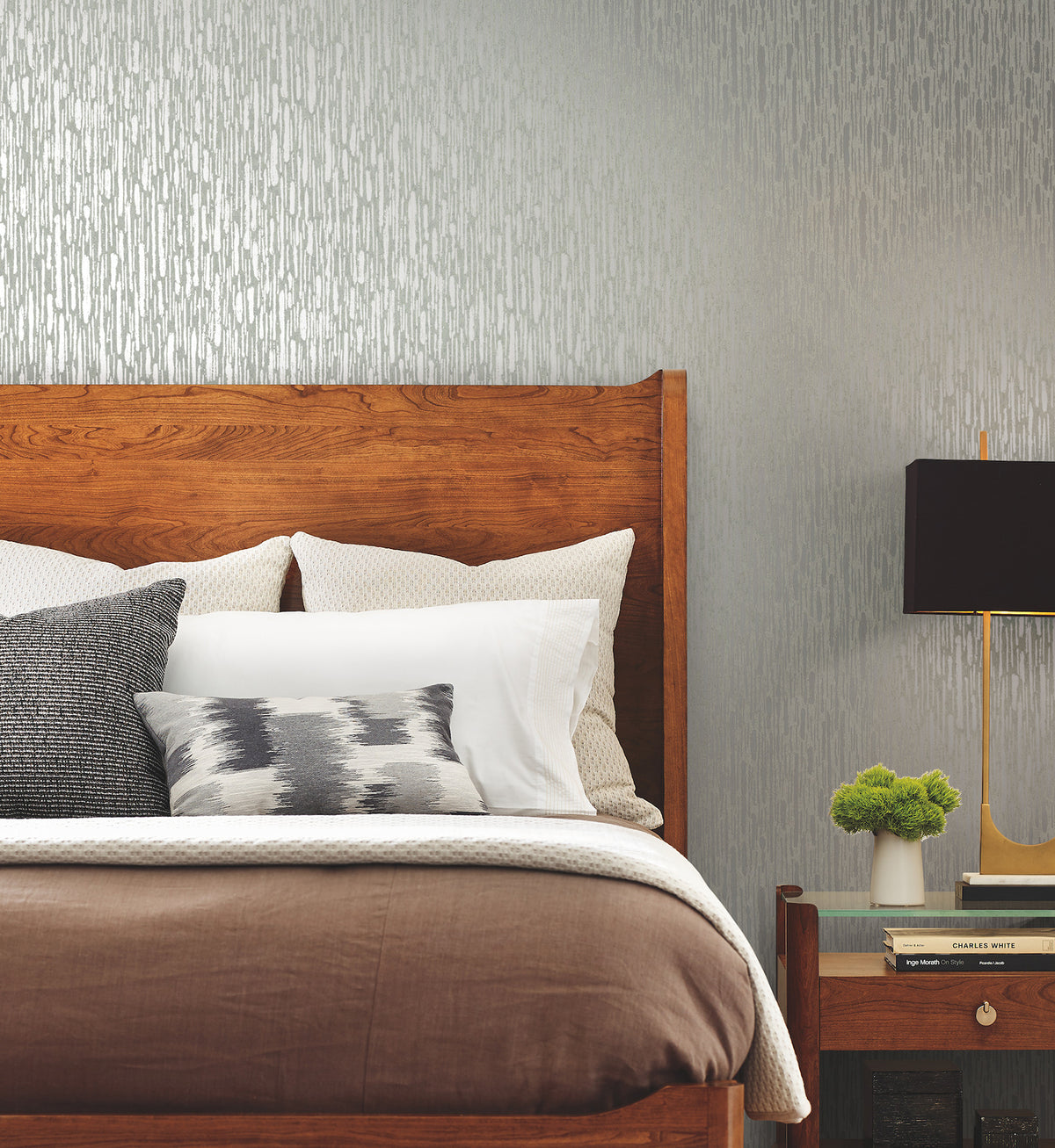 A wooden bed with white and gray pillows, a brown blanket, and a nightstand holding books, a lamp, and a small green plant stands against a wall adorned with York Wallcoverings’ Metallic Cascade Wallpaper.