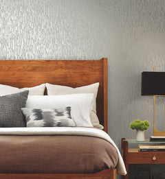 A neatly made bed with a wooden headboard, stacked pillows, and a brown and white color scheme sits next to a wooden nightstand topped with books, a lamp, and a small plant. The wall features York Wallcoverings’ Metallic Cascade Wallpaper, adding textured metallic elegance to the room.