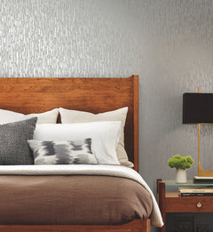 A modern bedroom featuring a wooden bed adorned with white and gray pillows and brown bedding, a nightstand displaying books, a small plant, and a black lamp. The wall is enhanced with York Wallcoverings’ Metallic Cascade Wallpaper in a textured silver-gray pattern.