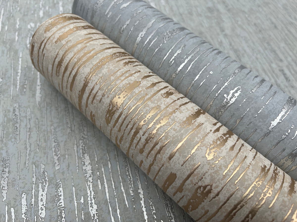 Two rolls of York Wallcoverings Metallic Cascade Wallpaper—one featuring a gold and beige design, the other showcasing a silver and gray pattern—are displayed on a coordinating textured surface.
