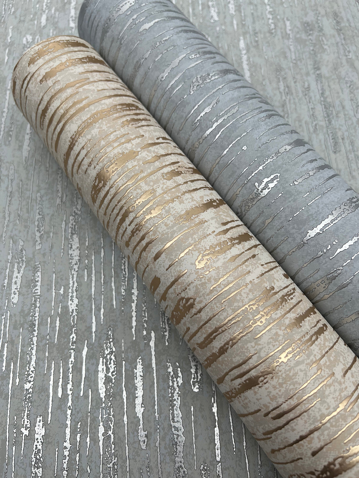Two rolls of York Wallcoverings’ Metallic Cascade Wallpaper, one featuring gold tones and the other silver, display a birch tree bark pattern as they rest on a coordinating textured surface.