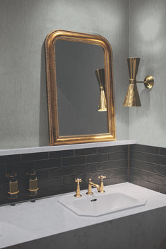 A bathroom vanity features a gold faucet, black tiled backsplash, gold-framed mirror, and a gold wall sconce. Two soap dispensers are mounted on the wall above the white countertop. The light gray wall is adorned with Metallic Cascade Wallpaper by York Wallcoverings.