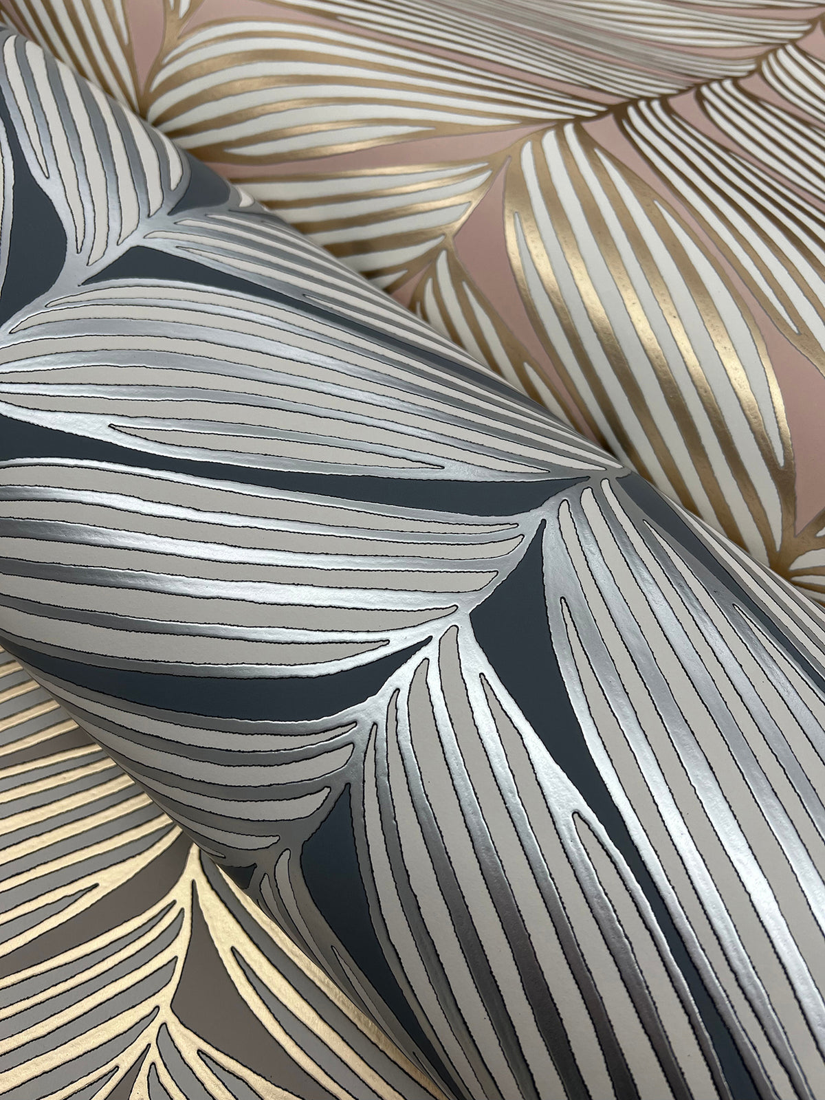 Close-up of York Wallcoverings’ Luminous Leaves Wallpaper, showcasing metallic gold and silver leaf patterns on pink and dark gray backgrounds. The design features overlapping, stylized leaves with fine, shimmering lines.