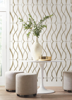 A round white table with a white vase holding green branches and a gold tray, surrounded by beige ottomans, sits in front of a wall adorned with York Wallcoverings’ Graceful Geo Wallpaper.
