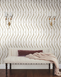 A white tufted bench with a mauve pillow and beige throw sits against a wall adorned with York Wallcoverings’ Graceful Geo Wallpaper, flanked by two gold and white wall sconces on a dark wood floor.