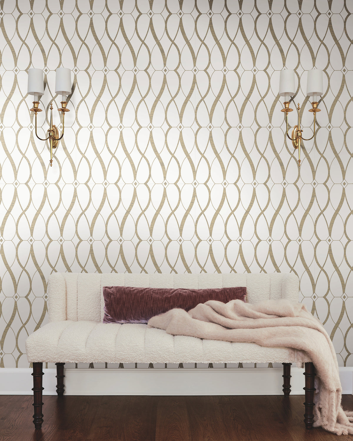 A wall adorned with York Wallcoverings’ Graceful Geo Wallpaper in a cream and gold pattern features two wall sconces above a cream upholstered bench. A mauve pillow and a soft beige throw blanket rest on the bench, which sits atop a dark wood floor.