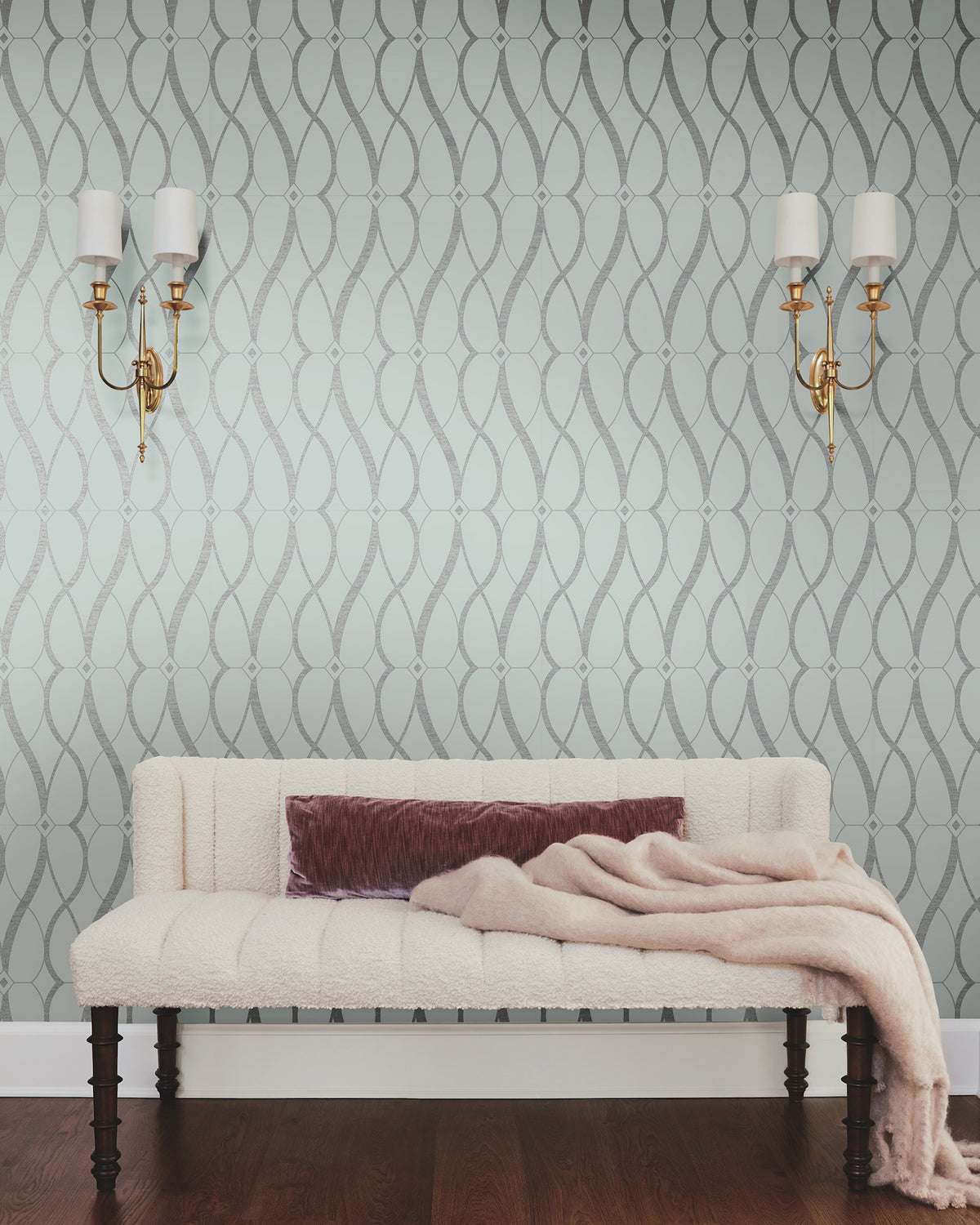 A cream bench with a mauve pillow and blanket sits against a wall adorned with Graceful Geo Wallpaper by York Wallcoverings. Two gold wall sconces with white shades hang on either side above the bench, while the floor is dark wood.