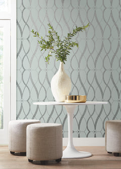 A white vase with green branches sits on a round white table next to a gold tray, surrounded by three beige stools. The backdrop features York Wallcoverings’ Graceful Geo Wallpaper and a bright window on the left.