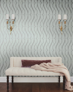 A cream-colored bench with dark wooden legs is topped with a mauve pillow and a soft beige throw. In the background, the walls are adorned with *Graceful Geo Wallpaper* by York Wallcoverings and flanked by two gold wall sconces with white shades.
