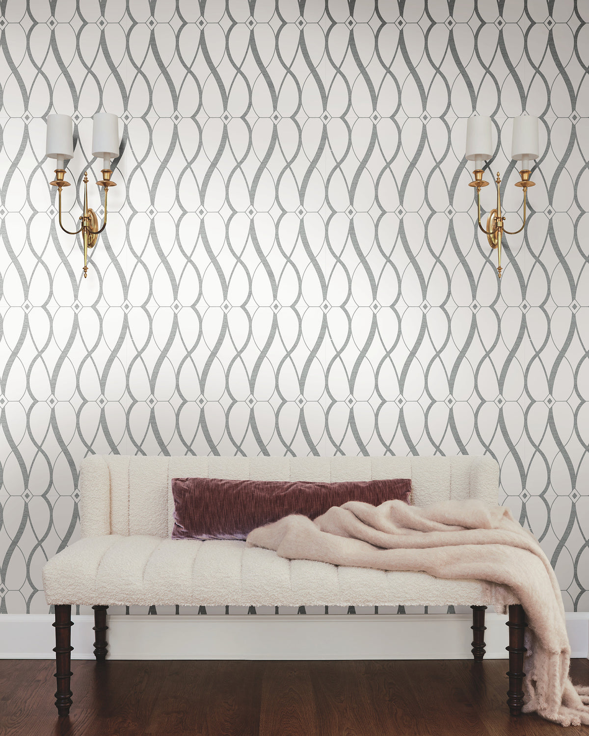 A cream upholstered bench with a mauve pillow and beige throw sits against a wall adorned with York Wallcoverings’ Graceful Geo Wallpaper, flanked by two gold wall sconces with white shades, on a dark wood floor.