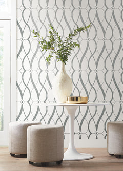 A white round table with a white vase of green branches and a gold tray is surrounded by three beige ottomans, set against York Wallcoverings’ Graceful Geo Wallpaper.