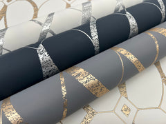 Three rolls of Graceful Geo Wallpaper by York Wallcoverings, featuring metallic geometric patterns in silver and gold on dark gray, light gray, and white backgrounds, displayed on a matching patterned surface.