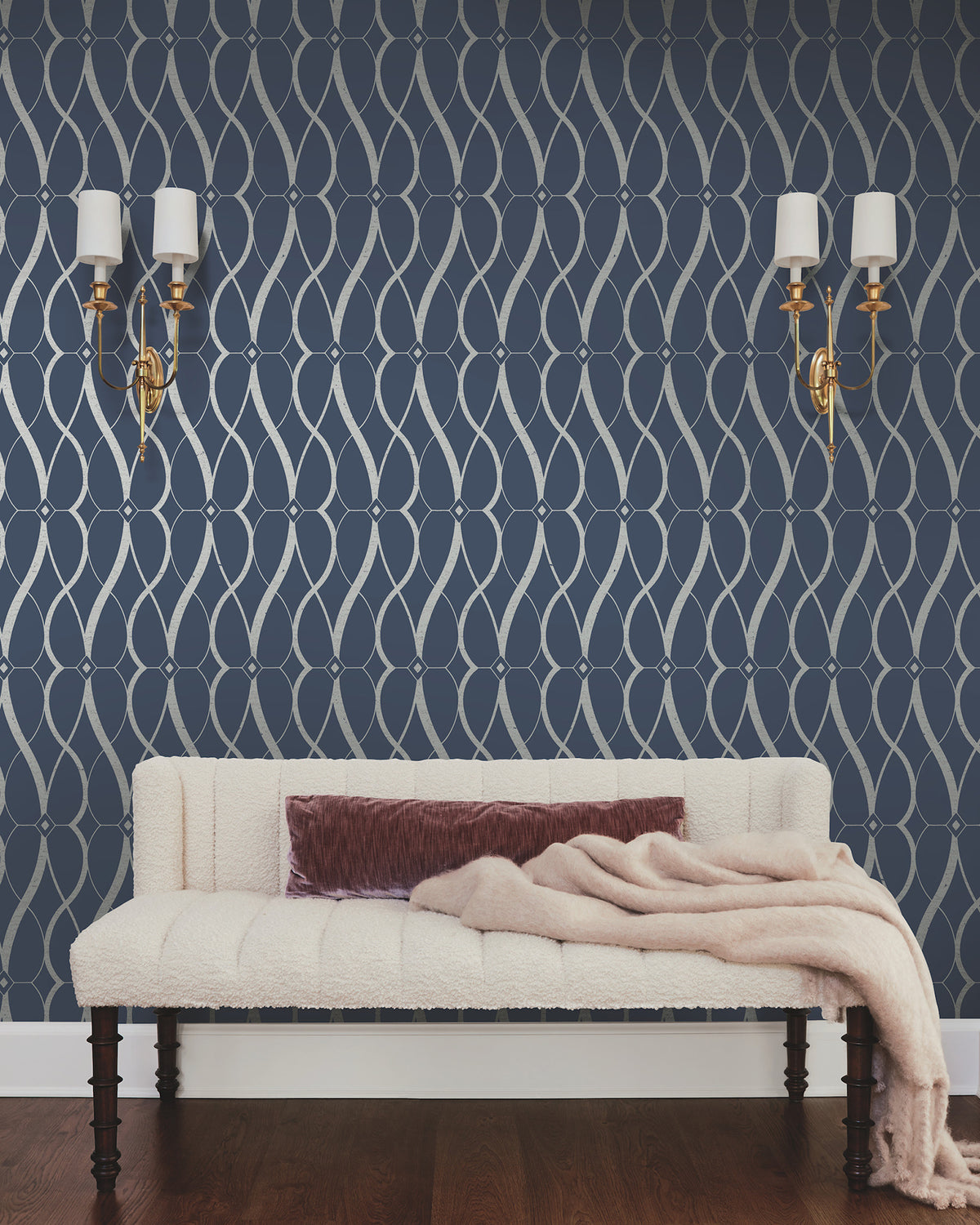 A cream-colored couch with a mauve pillow and a light pink throw sits against a navy wall adorned with York Wallcoverings’ Graceful Geo Wallpaper in a silver geometric pattern. Two gold wall sconces with white shades are mounted above the couch.