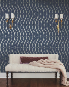 A cream-colored couch with a mauve pillow and a light pink throw sits against a navy wall adorned with York Wallcoverings’ Graceful Geo Wallpaper in a silver geometric pattern. Two gold wall sconces with white shades are mounted above the couch.