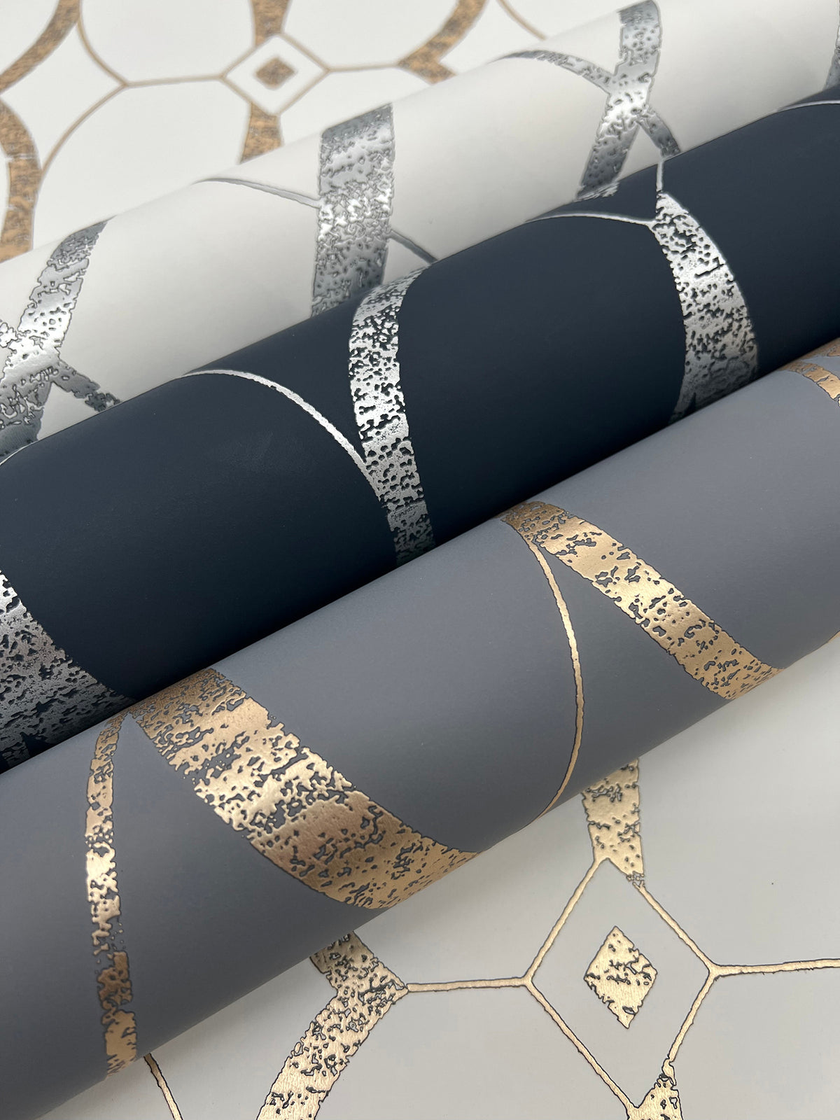 Three rolls of Graceful Geo Wallpaper by York Wallcoverings—in white with silver, black with silver, and gray with gold metallic geometric patterns—are displayed on a patterned surface that matches the wallpaper designs.