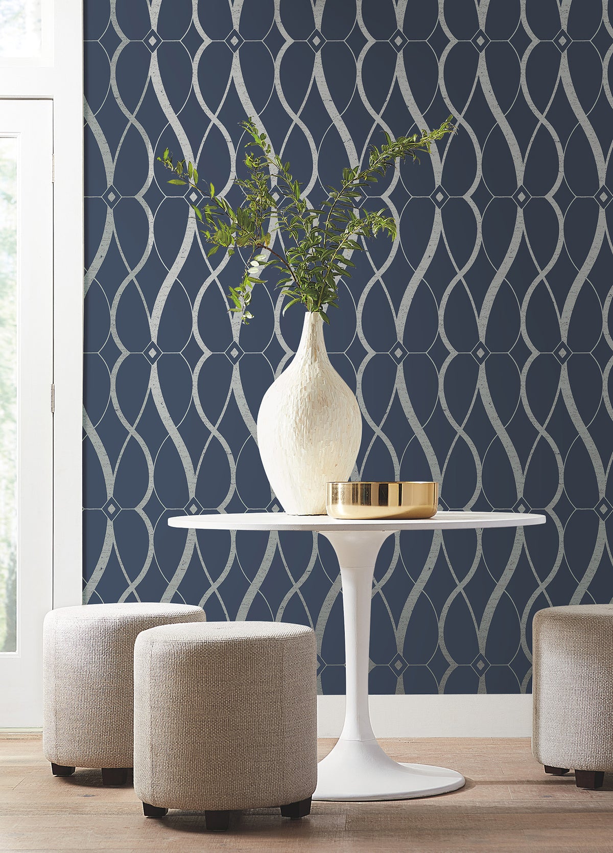 A modern round white table topped with a white vase of greenery sits between three beige ottomans, set against a dark blue wall adorned with York Wallcoverings’ Graceful Geo Wallpaper in a silver geometric pattern. Sunlight streams in through a window on the left.