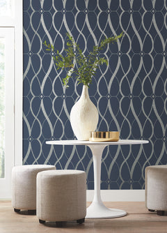 A modern round white table topped with a white vase of greenery sits between three beige ottomans, set against a dark blue wall adorned with York Wallcoverings’ Graceful Geo Wallpaper in a silver geometric pattern. Sunlight streams in through a window on the left.