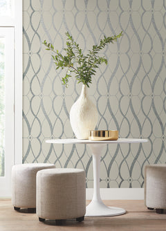 A small white round table with a tall white vase holding green branches, a gold tray, and two beige ottomans is set in front of York Wallcoverings’ Graceful Geo Wallpaper. Bright natural light enters from the left.