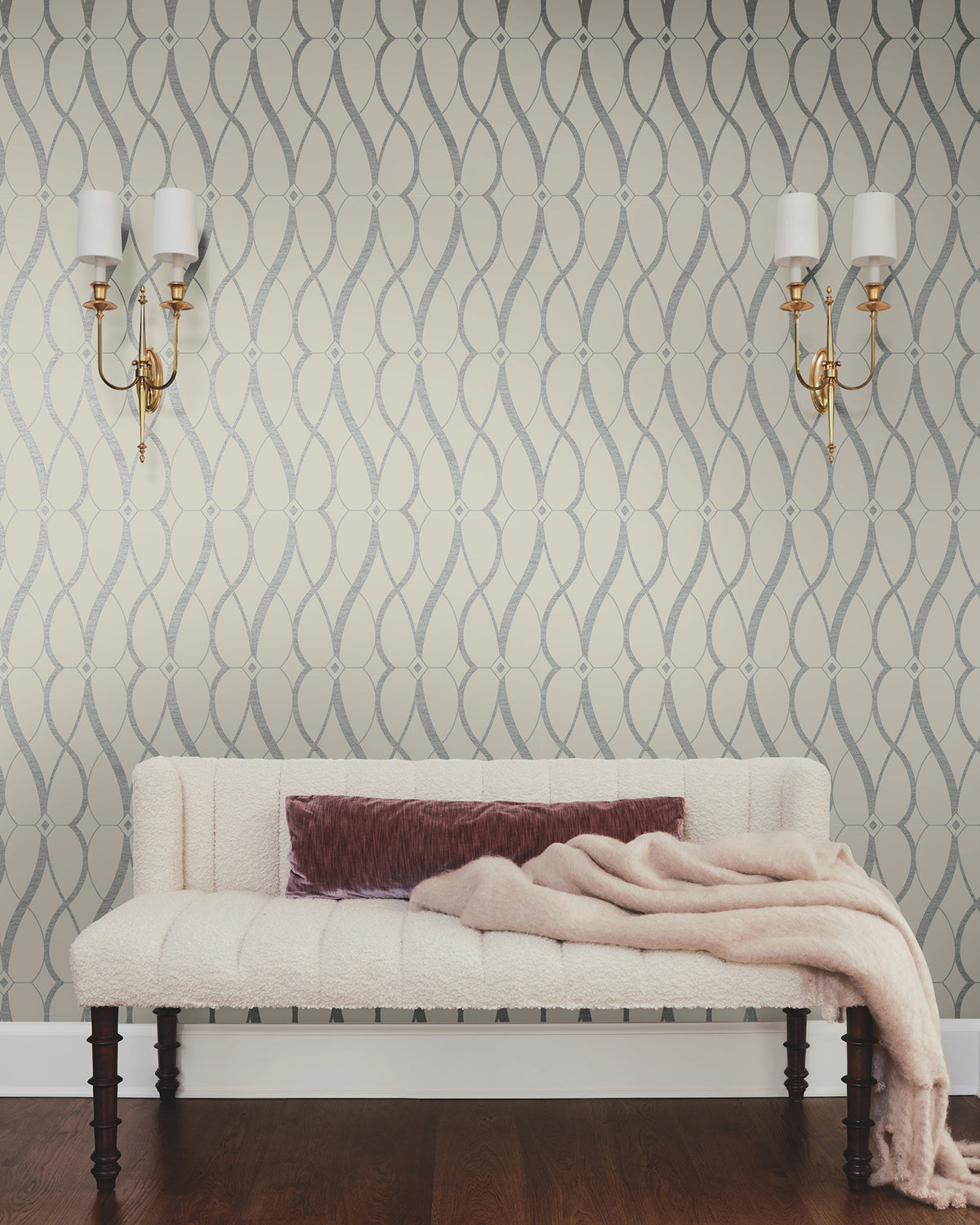 A cream-colored tufted sofa with a mauve pillow and light pink throw sits against the Graceful Geo Wallpaper by York Wallcoverings. Two brass wall sconces with white shades are mounted above the sofa on either side.