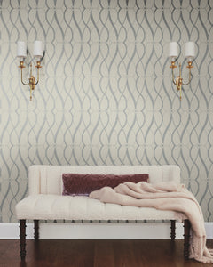 A cream-colored couch with a mauve pillow and beige throw blanket sits against a wall adorned with Graceful Geo Wallpaper by York Wallcoverings. Two gold wall sconces with white shades are mounted above the couch, and dark wood flooring is visible.