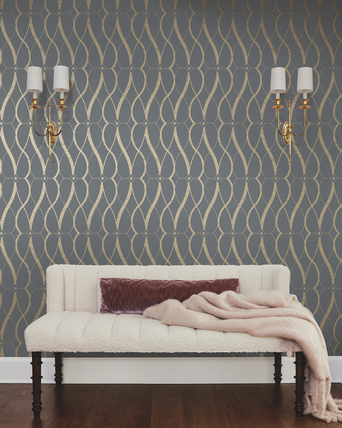 A cream-colored sofa with a mauve throw blanket sits against a wall adorned with Graceful Geo Wallpaper by York Wallcoverings in a dark gray and gold geometric pattern. Two brass wall sconces with white shades are mounted above the sofa.