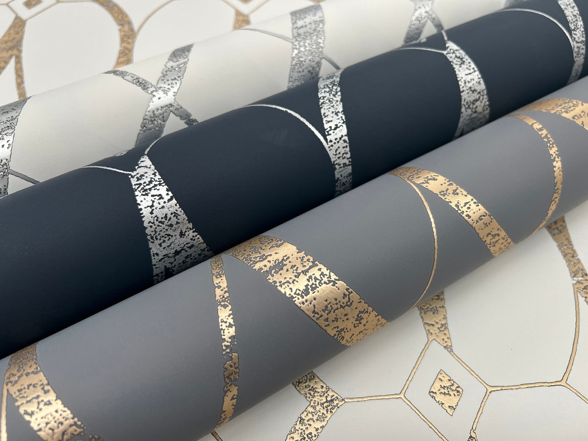 Three rolls of York Wallcoverings’ Graceful Geo Wallpaper, featuring metallic geometric patterns in silver and gold on dark blue, gray, and off-white backgrounds, are displayed on a matching patterned surface.