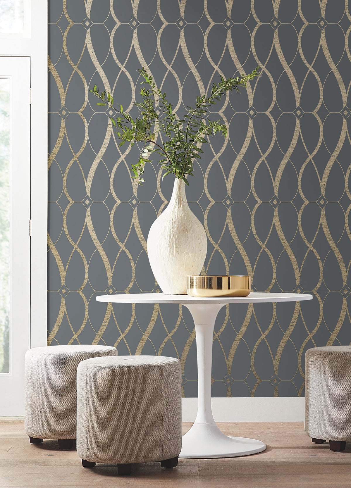 A white vase with green branches and a gold tray sit on a round white table, flanked by two beige stools, against a dark wall covered in Graceful Geo Wallpaper by York Wallcoverings. Light enters from a window on the left.