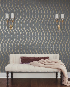 A cream-colored sofa with a mauve pillow and throw sits against a dark gray wall adorned with the Graceful Geo Wallpaper by York Wallcoverings. Two gold sconces with white shades are mounted above the sofa, and the floor is dark wood.