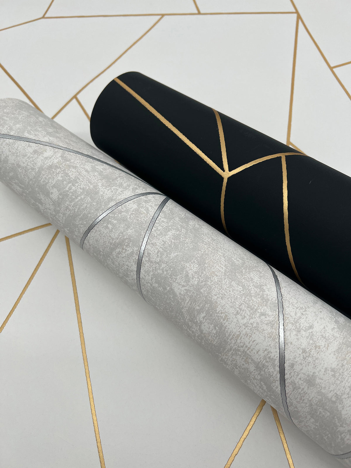 Two rolls of York Wallcoverings’ Nazca Wallpaper are displayed on a geometric-patterned surface: one roll is black with gold lines, the other is light gray with silver lines. The background also features intersecting gold lines.