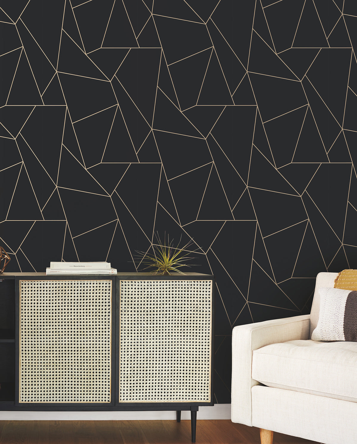A modern living room featuring the Nazca Wallpaper by York Wallcoverings—a striking geometric black wall—paired with a light-colored couch, patterned sideboard, and decorative accents such as a spiky gold sculpture and stacked books.