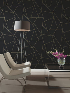 Two modern white chairs and a black lamp sit by a black coffee table with a glass vase of purple flowers, all set against York Wallcoverings’ Nazca Wallpaper in black with a gold geometric line pattern.