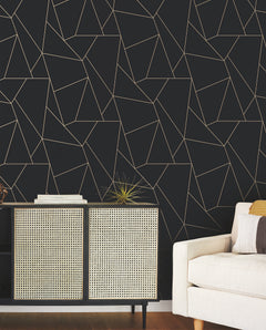 Modern living room with a beige sofa, a cabinet with rattan doors, a decorative plant, and a dark wall featuring the Nazca Wallpaper by York Wallcoverings with its geometric gold line pattern.