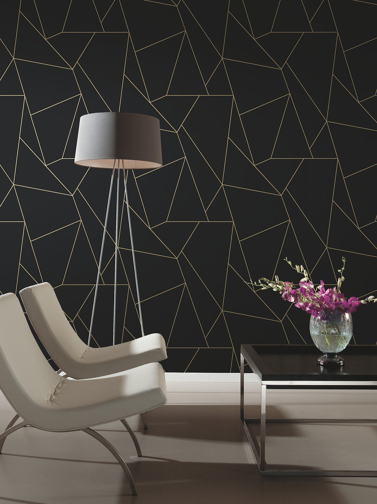 Two modern white chairs and a black floor lamp face a sleek black coffee table with a vase of purple flowers, set against a wall adorned with Nazca Wallpaper by York Wallcoverings, featuring a thin gold geometric pattern.