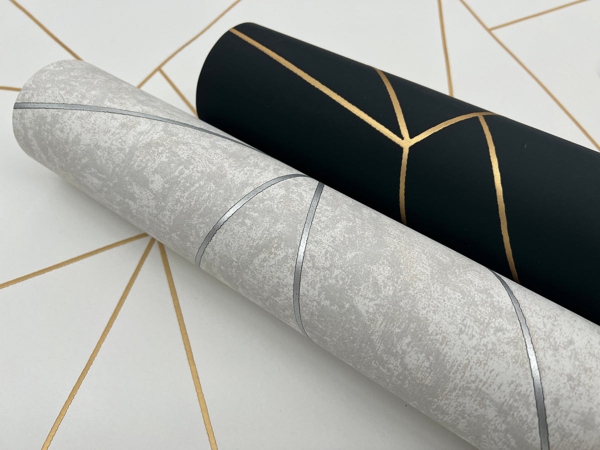 Two rolls of York Wallcoverings’ Nazca Wallpaper are displayed on a white surface; one roll features a light gray background with silver geometric lines, while the other showcases a black background with gold geometric patterns.