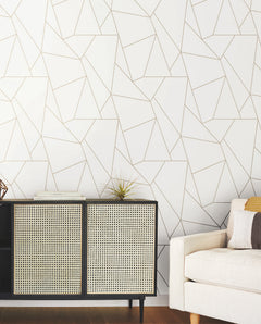 A modern living room with a beige sofa, a black and tan cabinet topped with a small plant and books, and a white wall adorned with the “Nazca Wallpaper” by York Wallcoverings featuring a gold geometric line pattern.