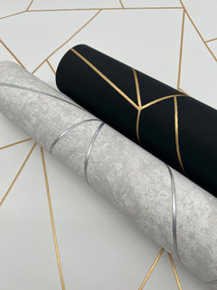 Two rolls of Nazca Wallpaper by York Wallcoverings—one in black with gold geometric lines and the other in light gray with silver lines—are displayed on a white surface adorned with gold geometric patterns.