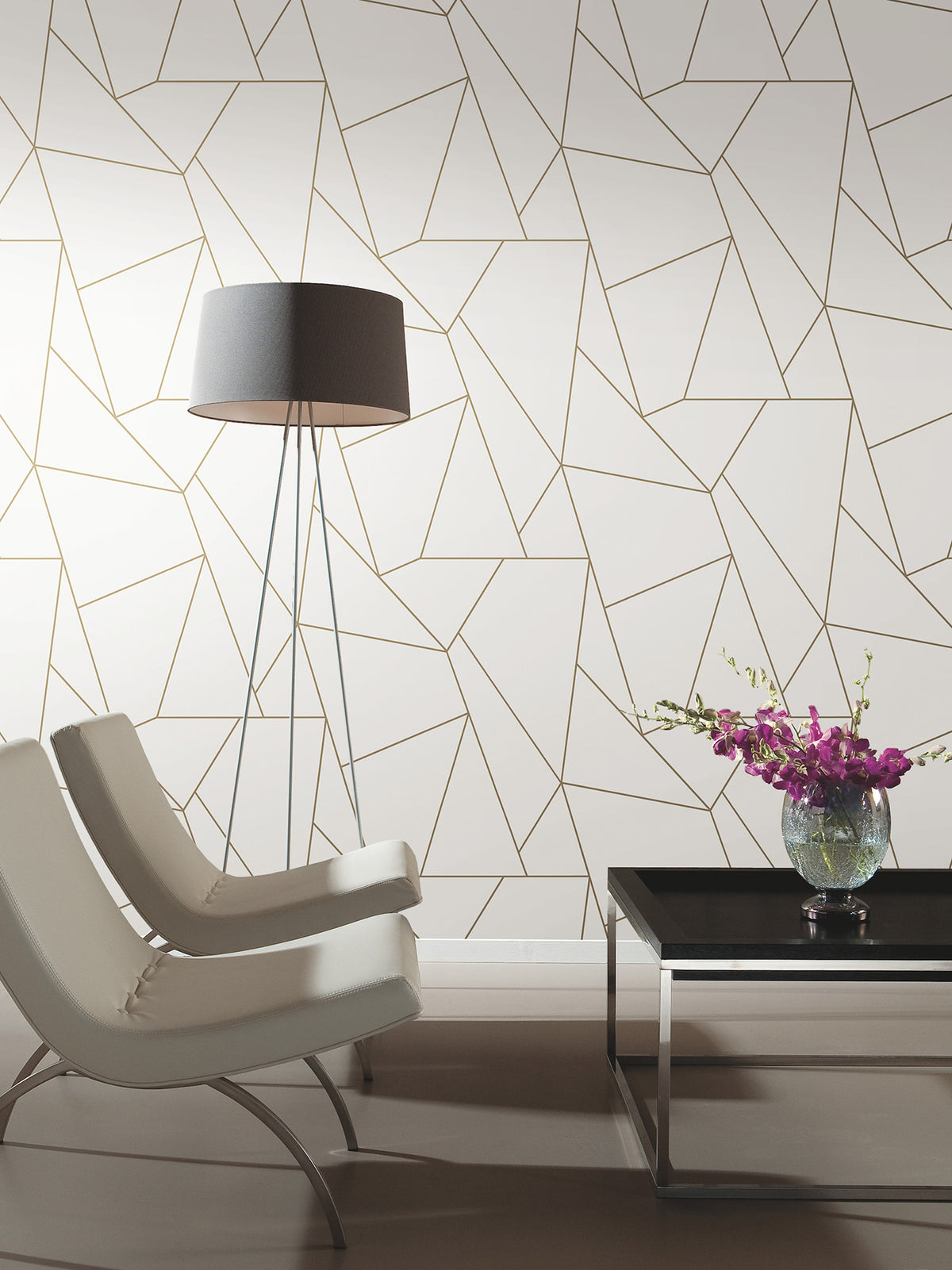 Two modern white chairs face a black coffee table with a glass vase of purple flowers. A tall floor lamp stands beside a wall covered in York Wallcoverings’ Nazca Wallpaper, featuring a geometric gold and white pattern.