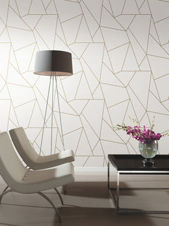 Two modern white chairs face a black coffee table with a glass vase of purple flowers. A tall floor lamp stands beside a wall covered in York Wallcoverings’ Nazca Wallpaper, featuring a geometric gold and white pattern.