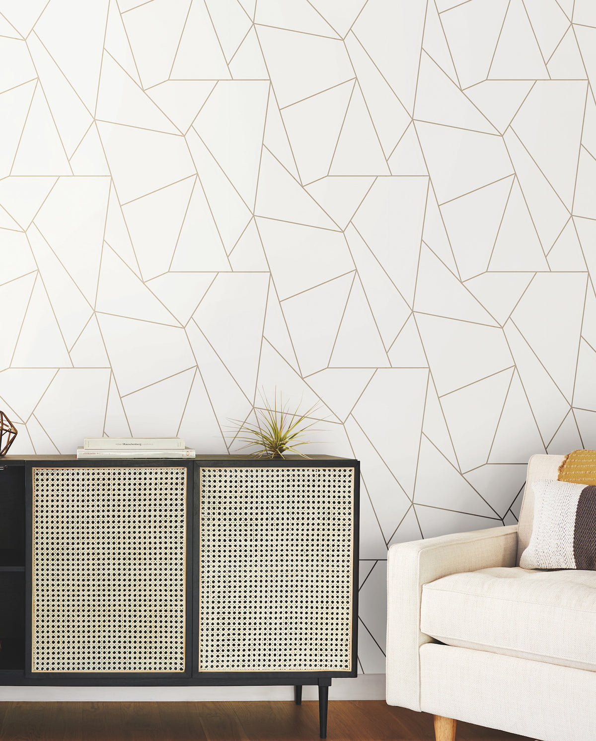 A modern room featuring a beige sofa and a black and wicker cabinet adorned with decor, set against a white wall covered in York Wallcoverings’ Nazca Wallpaper with a gold geometric line pattern. The flooring is wooden.