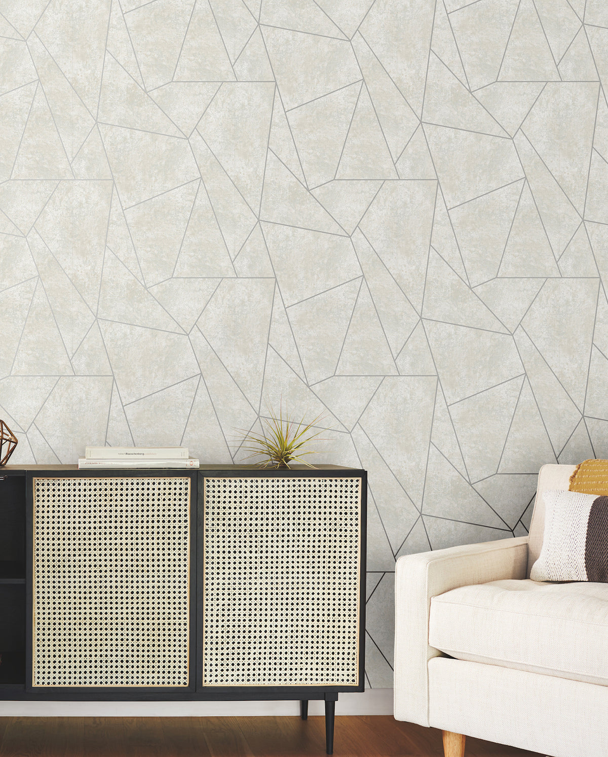 A modern living room featuring a beige sofa, a black and cane cabinet, books, a small plant, and York Wallcoverings’ Nazca Wallpaper in a light gray geometric pattern.