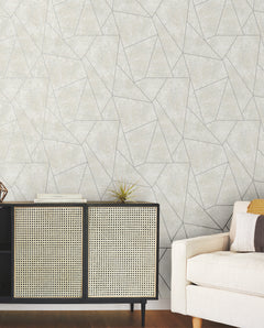 A modern living room featuring a beige sofa, a black and cane cabinet, books, a small plant, and York Wallcoverings’ Nazca Wallpaper in a light gray geometric pattern.