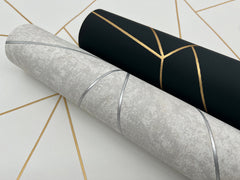 Two rolls of Nazca Wallpaper by York Wallcoverings are displayed on a white surface with gold geometric lines. One roll features a gray background with silver lines, while the other showcases a black background with gold geometric patterns.